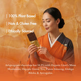 A woman in an orange and black traditional outfit holds a chocolate bar wrapped in gold foil against a warm orange textured background. White text on the image highlights that the product is 100% plant-based, nuts and gluten free, ethically sourced, and contains adaptogenic ingredients including Lion's Mane mushroom, green tea, ginseng, ginkgo biloba, and astragalus.