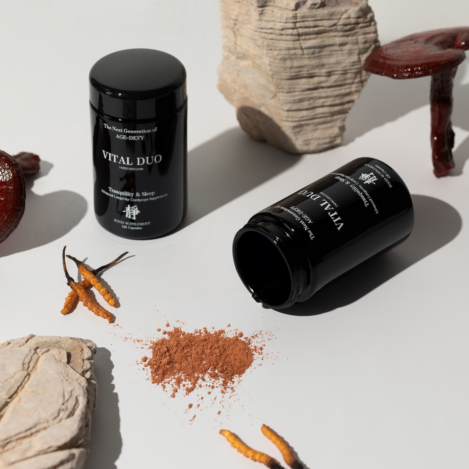 Two black cylindrical supplement jars labeled VITAL DUO are arranged on a white surface with dried orange herbs and rocks around them. One jar is upright and the other lies on its side with brown powder spilled out.