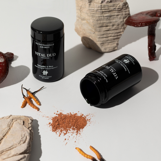 Two black cylindrical supplement jars labeled VITAL DUO are arranged on a white surface with dried orange herbs and rocks around them. One jar is upright and the other lies on its side with brown powder spilled out.