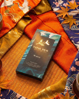A rectangular box of Vital Duo Water tea by Crafted Melodies lies on a woven mat, surrounded by colorful patterned fabrics in orange, blue, and gold.
